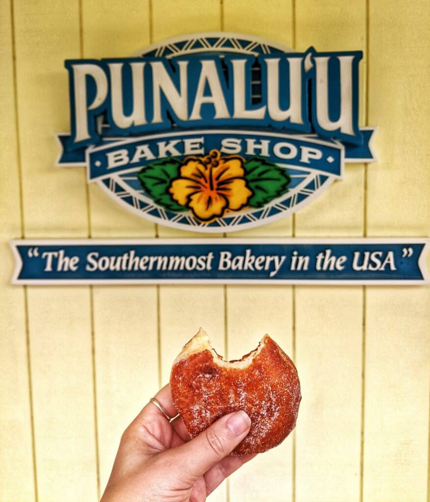 my hand holding a malasada with one bite taken out in front of the punaluu bake shop sign