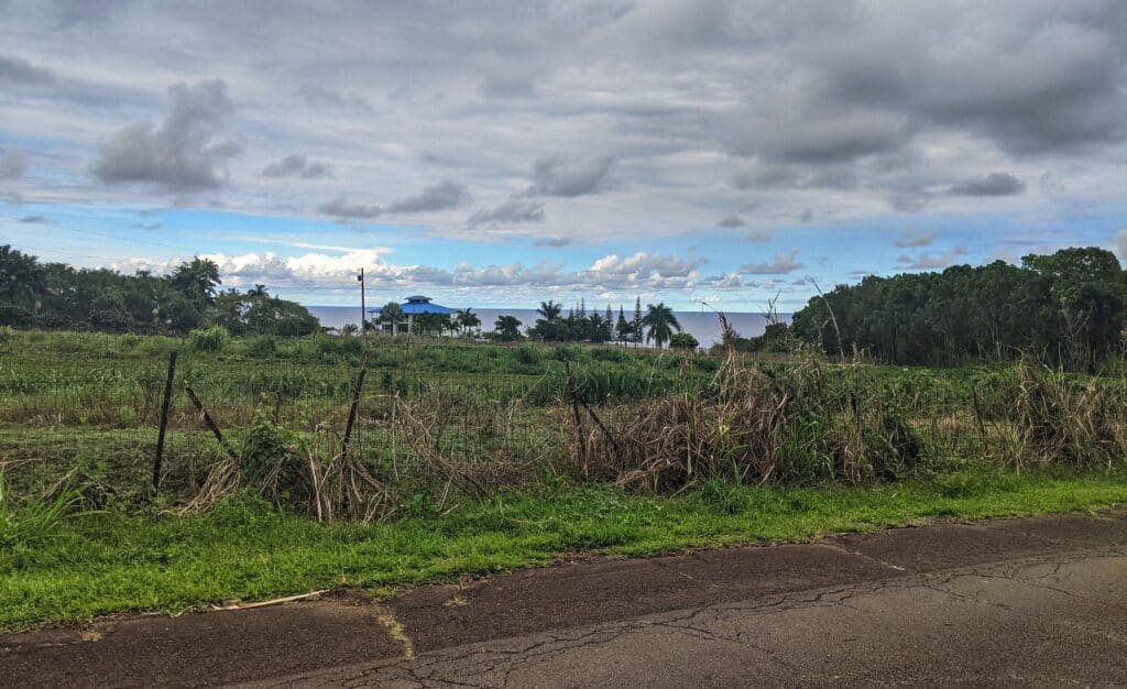 Rustic Hawaiian landscape with fields and fences leading towards the ocean, under a sky partly cloudy, partly blue
