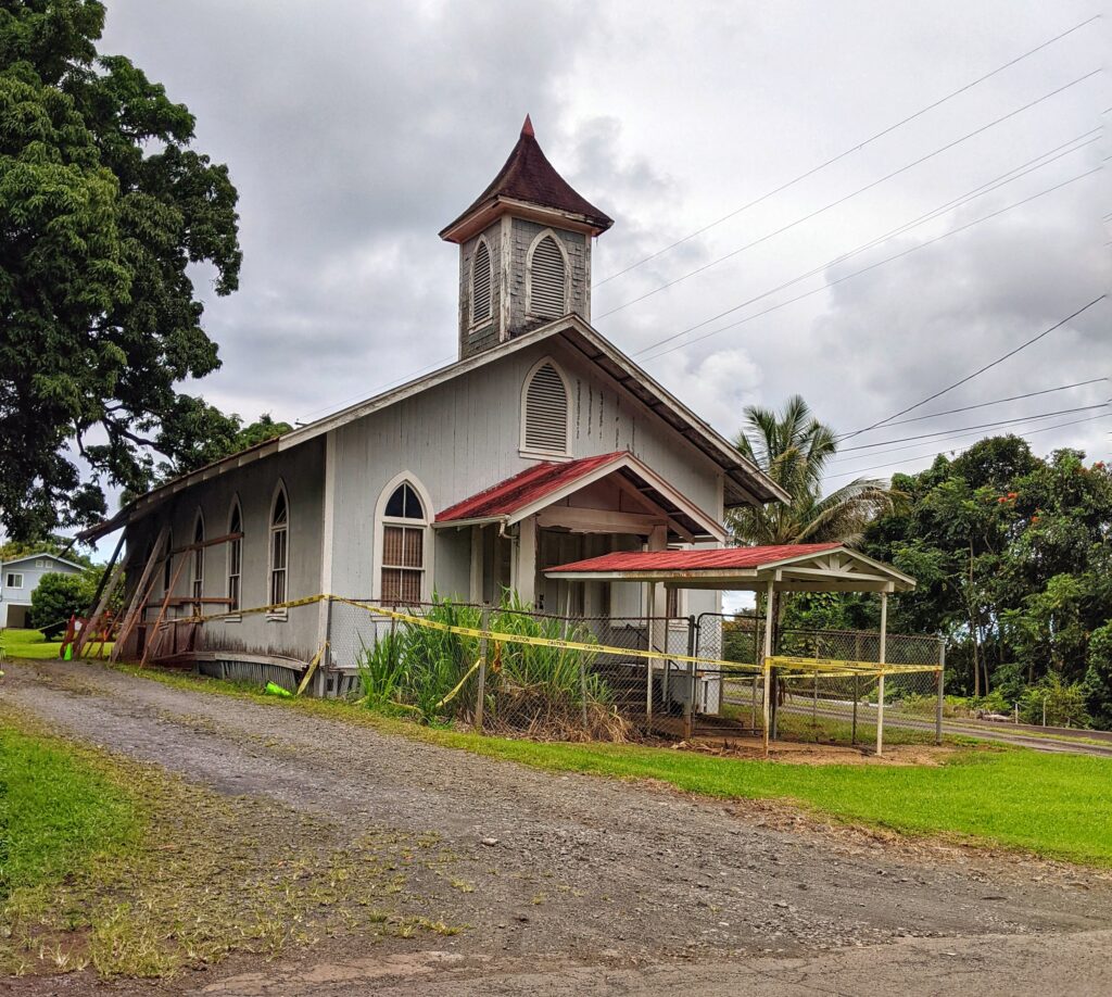 Historic abandoned wooden church with a faded red roof nestled among tropical foliage, showcasing the serene beauty of Pepe'ekeo's past.