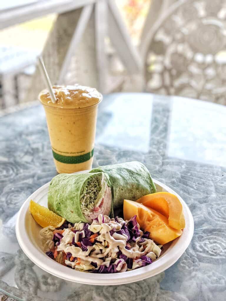 Healthy meal featuring a green wrap, orange slices, and coleslaw on a white plate, paired with a mango smoothie on a patterned glass tabletop.