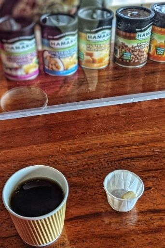 hamakua macadamia nut samples and A cup of black coffee in focus with blurred jars of local Hawaiian macadamia nuts in the background,