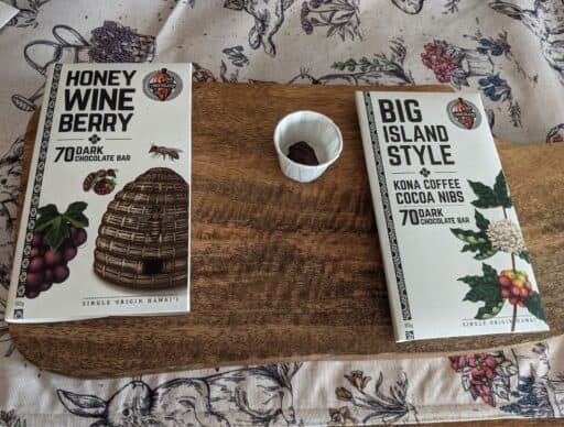 Two bars of Hawaiian chocolate, 'Honey Wine Berry' and 'Big Island Style Kona Coffee Cocoa Nibs', on a wooden table with a fabric cloth
