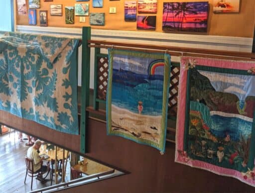 Quilts with Hawaiian themes, including sea life and landscapes, displayed over a railing above a café setting