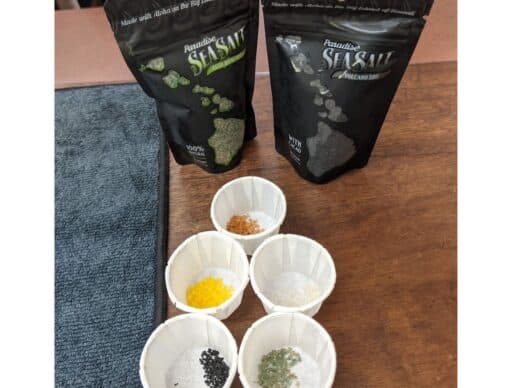 Assorted Hawaiian sea salts in tasting cups, with packets of Paradise Sea Salt in the background, on a wooden table. these make good hawaiian souvenirs