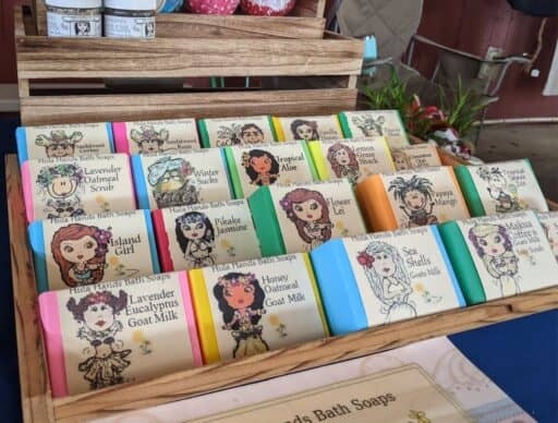 A variety of scented handmade soaps by Hula Hands Bath Soaps with playful illustrations and names like 'Lavender Oatmeal Scrub' and 'Pikake Jasmine'.
