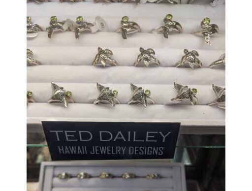 Silver rings with green peridot gemstones on display, labeled 'Ted Dailey Hawaii Jewelry Designs', set against a white cushioned background.