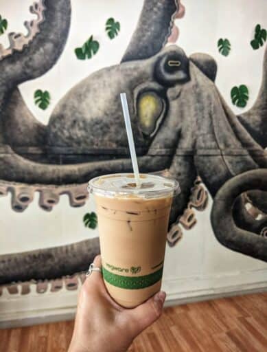 my hand holding a cup of honua's iced coffee with octopus mural in background