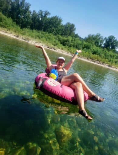 Woman relaxes with arms raised while floating the Wenatchee River in Leavenworth on a pink inner tube over clear water with visible rocks below. Green trees line the riverbank under a bright blue sky creating a fun and scenic summer float.