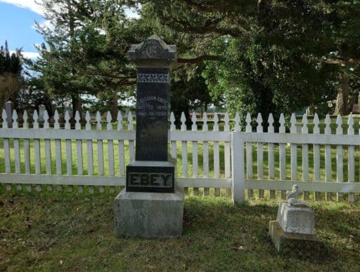 Historic gravestone marked "Ebey" stands beside a white picket fence and tall trees in a quiet cemetery near Coupeville. Visiting historic landmarks connected to Ebey’s Landing is one of the meaningful things to do in Coupeville.