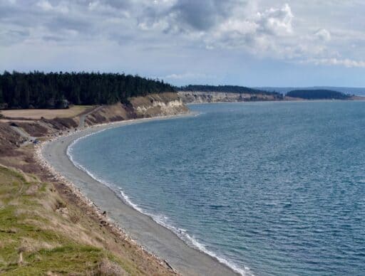 Curving pebble beach and rugged coastal cliffs near Coupeville with calm blue water and dramatic clouds overhead. Exploring scenic shorelines and hiking along Ebey’s Landing are unforgettable things to do in Coupeville.