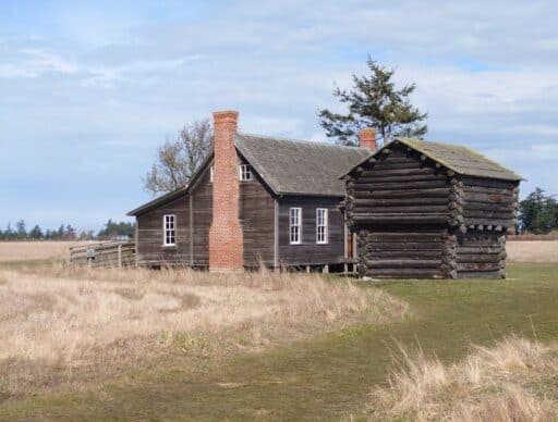 Historic wooden farmhouse and log cabin sitting in an open field near Coupeville under a cloudy sky. Visiting preserved pioneer homesteads and learning about early island life are interesting things to do in Coupeville