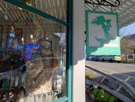 Boutique storefront in Coupeville with turquoise trim, a hanging "Aqua Gifts" sign featuring a mermaid silhouette, and colorful mermaid themed decor displayed in the window. Shopping for coastal gifts and local art is one of the charming things to do in Coupeville.
