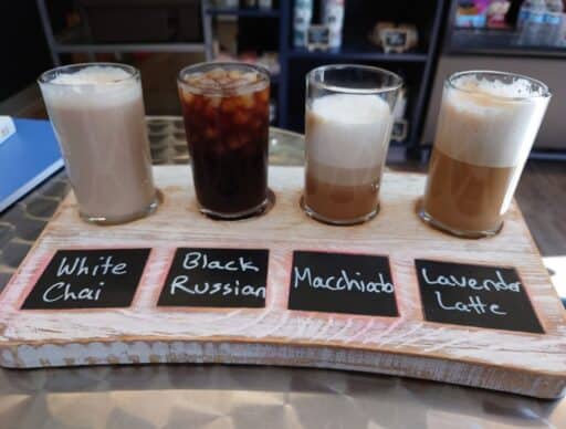 Four specialty coffee drinks sit on a wooden tasting board inside a cafe in Coupeville. Small chalkboard labels read "White Chai," "Black Russian," "Macchiato," and "Lavender Latte," highlighting a cozy coffee stop among the things to do in Coupeville.