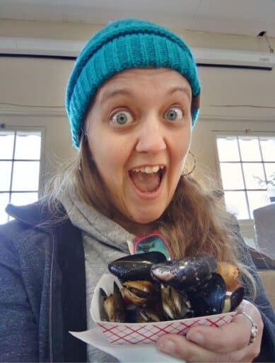 Smiling woman in a blue knit hat holds a tray of fresh mussels indoors in Coupeville. Sampling local Penn Cove mussels and fresh seafood is one of the most popular things to do in Coupeville.