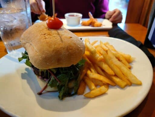 Burger topped with greens and a cherry tomato served with fries at a restaurant in Coupeville, with seafood appetizers visible in the background. Trying local restaurants and waterfront dining is one of the delicious things to do in Coupeville.