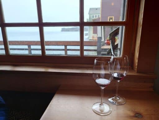 Two glasses of red wine sit on a wooden table beside a window overlooking the water in Coupeville. Enjoying waterfront dining and cozy views of Penn Cove is one of the relaxing things to do in Coupeville.