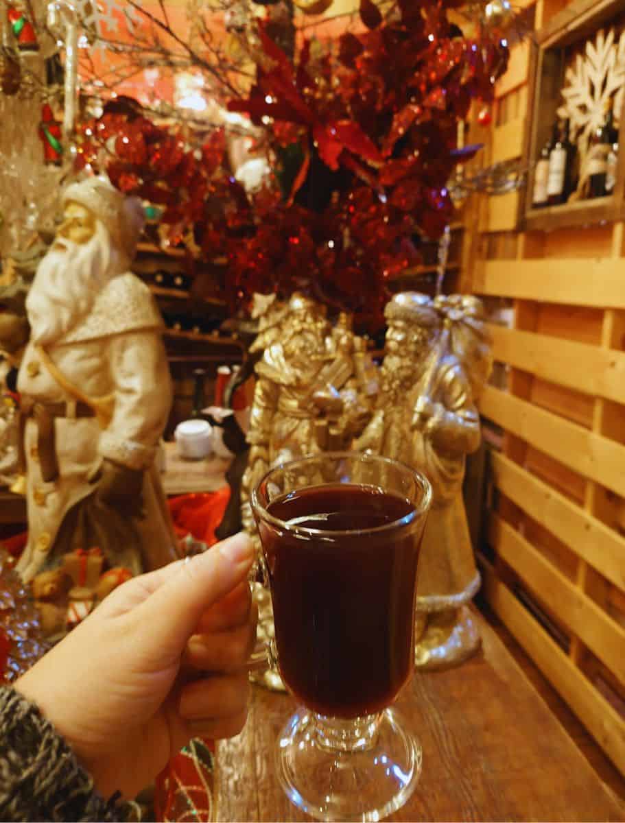 my hand holding a clear mug of gluhwein with christmas decor and statues in background