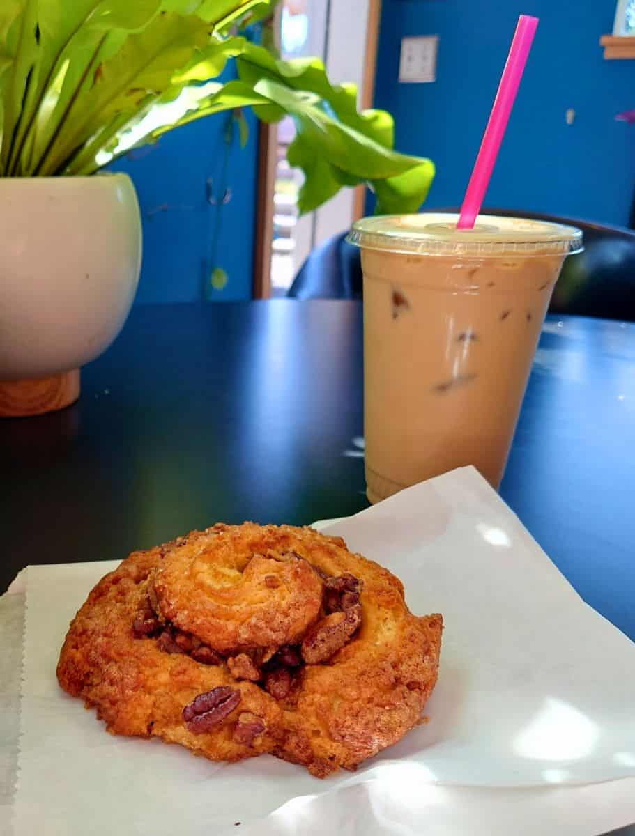 Iced latte with a bright pink straw beside a large cinnamon pastry topped with pecans on a café table inside a colorful coffee shop on Whidbey Island. Fresh pastries and specialty espresso drinks are part of what makes these cafés some of the best coffee spots on Whidbey Island.
