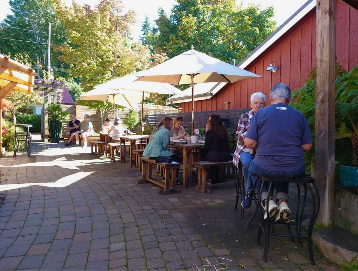 Outdoor patio seating area at a Whidbey Island coffee shop with people chatting at wooden tables beneath large umbrellas on a sunny day. Relaxing with coffee in welcoming outdoor spaces is part of discovering the best coffee on Whidbey Island.
