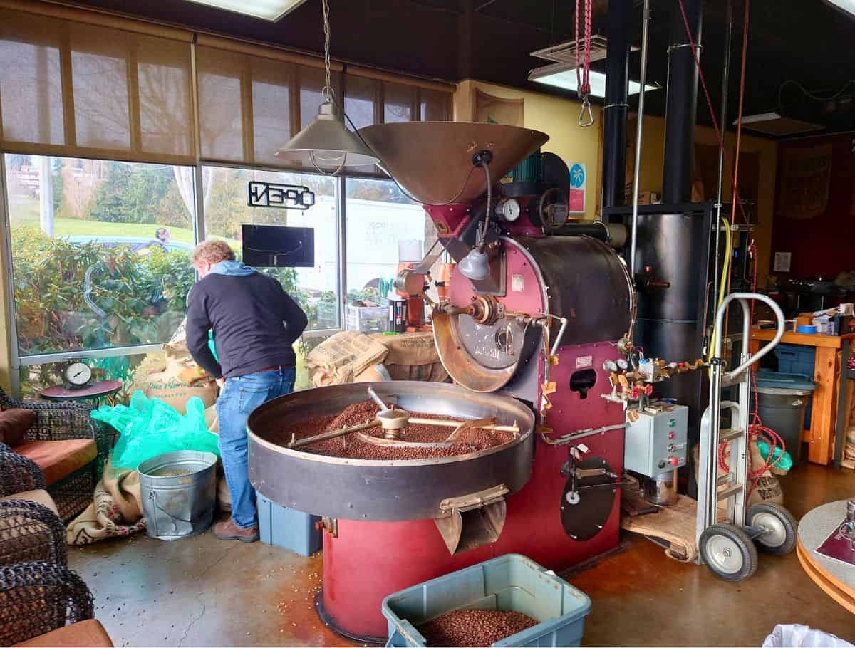Large coffee roasting machine filled with fresh beans inside a Whidbey Island coffee shop as a worker prepares bags of coffee nearby. Watching beans roast in house adds to the experience of finding the best coffee on Whidbey Island.