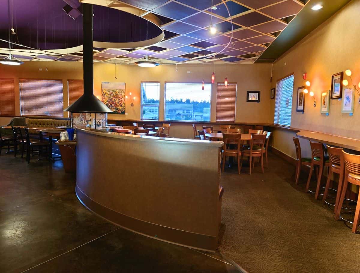Spacious coffee shop interior with warm lighting, wooden tables, cushioned seating, and a modern fireplace in the center of the room. Comfortable cafés like this make it easy to relax while exploring the best coffee on Whidbey Island.