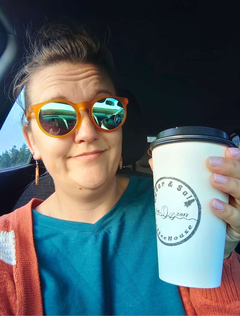Woman wearing orange sunglasses and a teal shirt holds a takeaway coffee cup from “Cedar and Salt Coffeehouse Est. 2017” while sitting in a car. Sampling local cafés like this is part of discovering the best coffee on Whidbey Island.