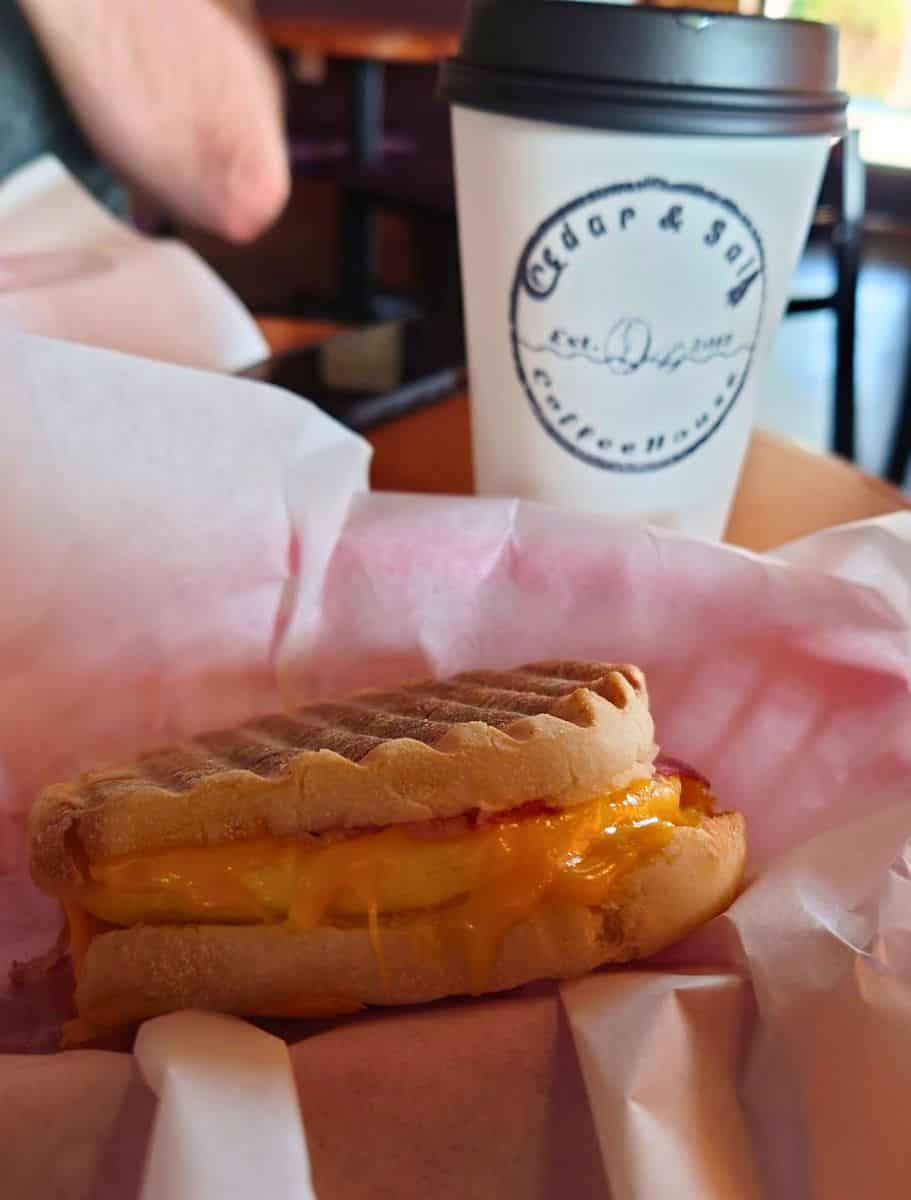 Breakfast sandwich with melted cheddar cheese on toasted bread served in a paper basket beside a takeaway cup from “Cedar and Salt Coffeehouse.” Cozy café breakfasts like this pair perfectly with some of the best coffee on Whidbey Island.