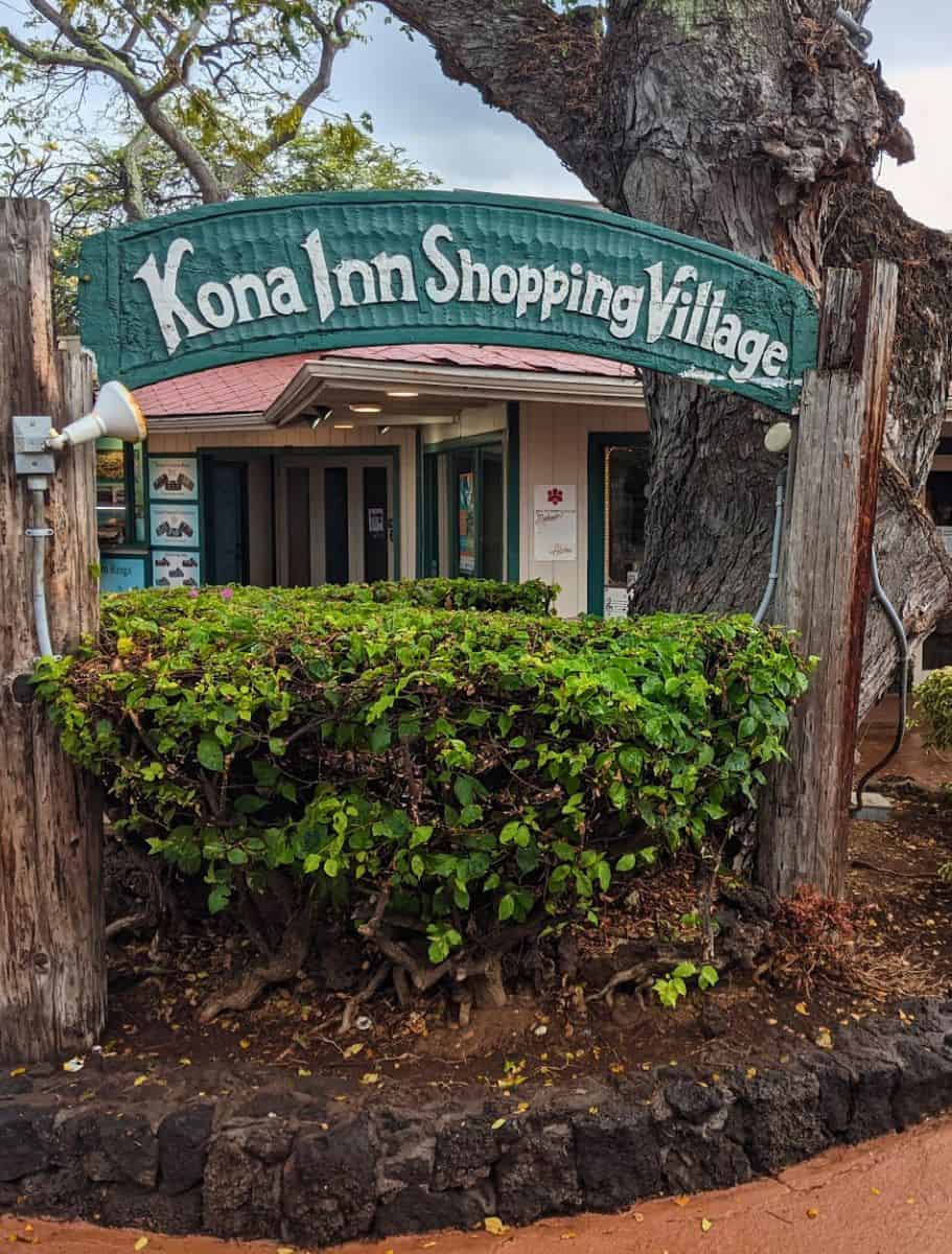 The rustic sign of Kona Inn Shopping Village nestled among greenery, representing shopping destinations that offer shelter and local goods during the rain