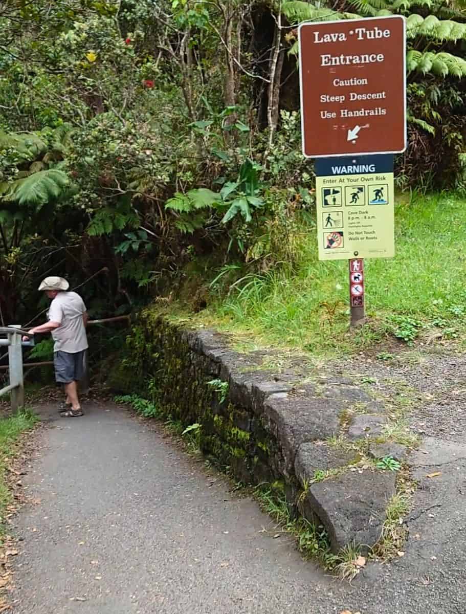 The entrance to the Thurston Lava Tube marked by a warning sign about the steep descent, as a visitor carefully holds the handrail beginning their journey.