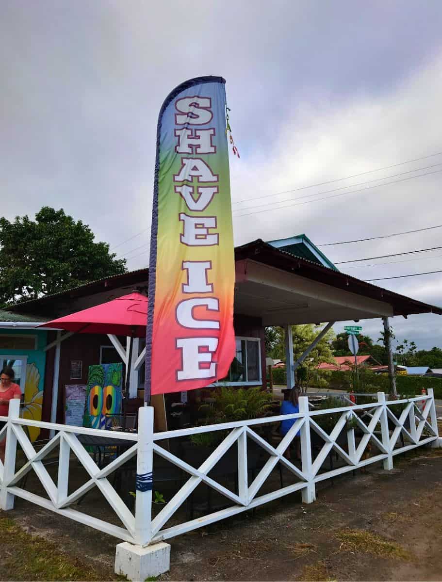 Colorful vertical banner spelling 'Shave Ice' towers above a quaint roadside stand with a white picket fence, inviting travelers on the Pepe'ekeo Scenic Drive to enjoy a local Hawaiian treat.