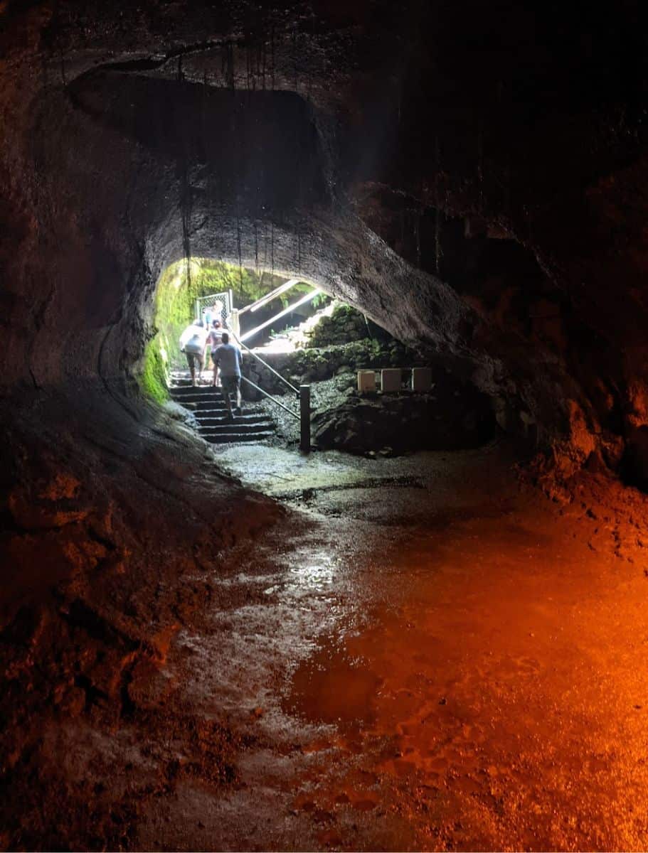Two visitors ascend a lit staircase in the Thurston Lava Tube, a contrast of light and shadow highlighting the cave's rugged interior