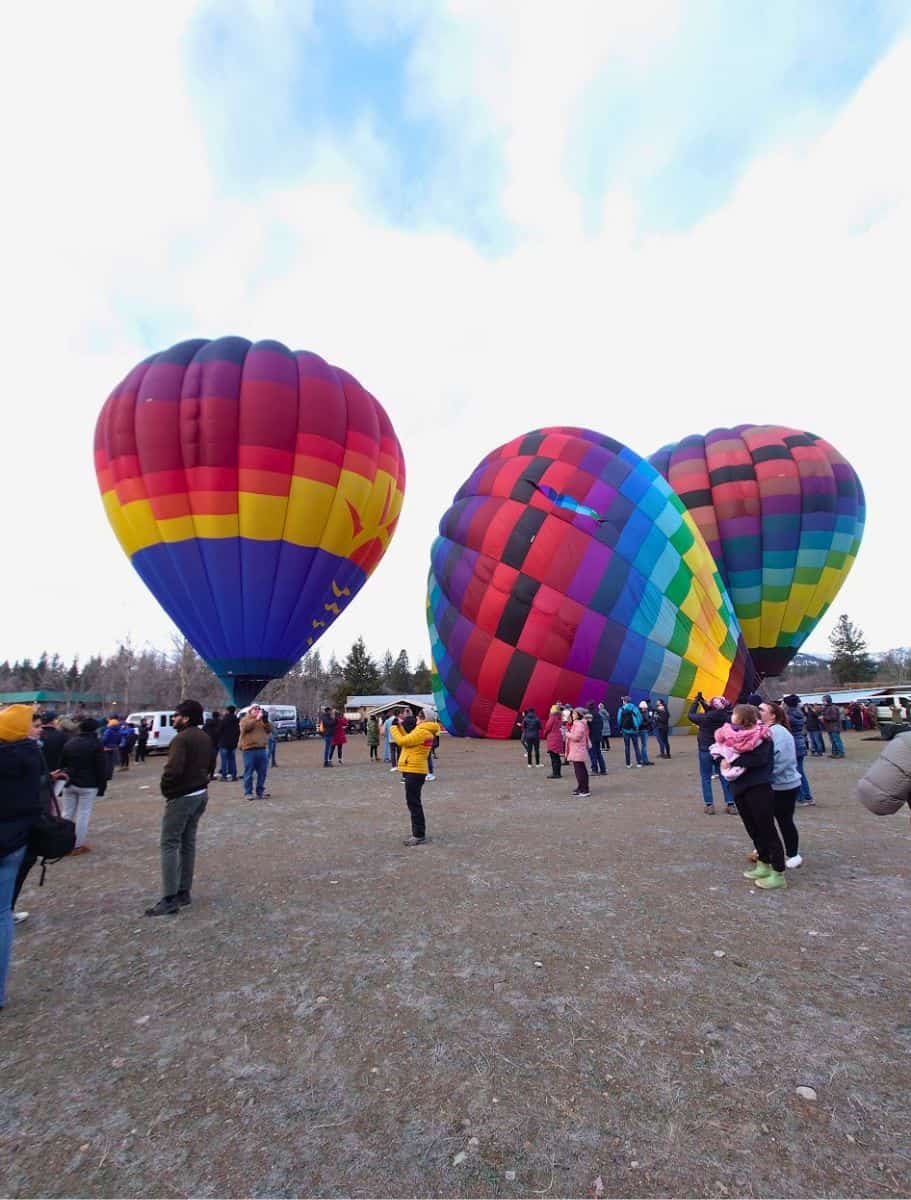 Three large, colorful hot air balloons fully inflated on a field, with a crowd of people admiring them against a backdrop of a cloudy sky
