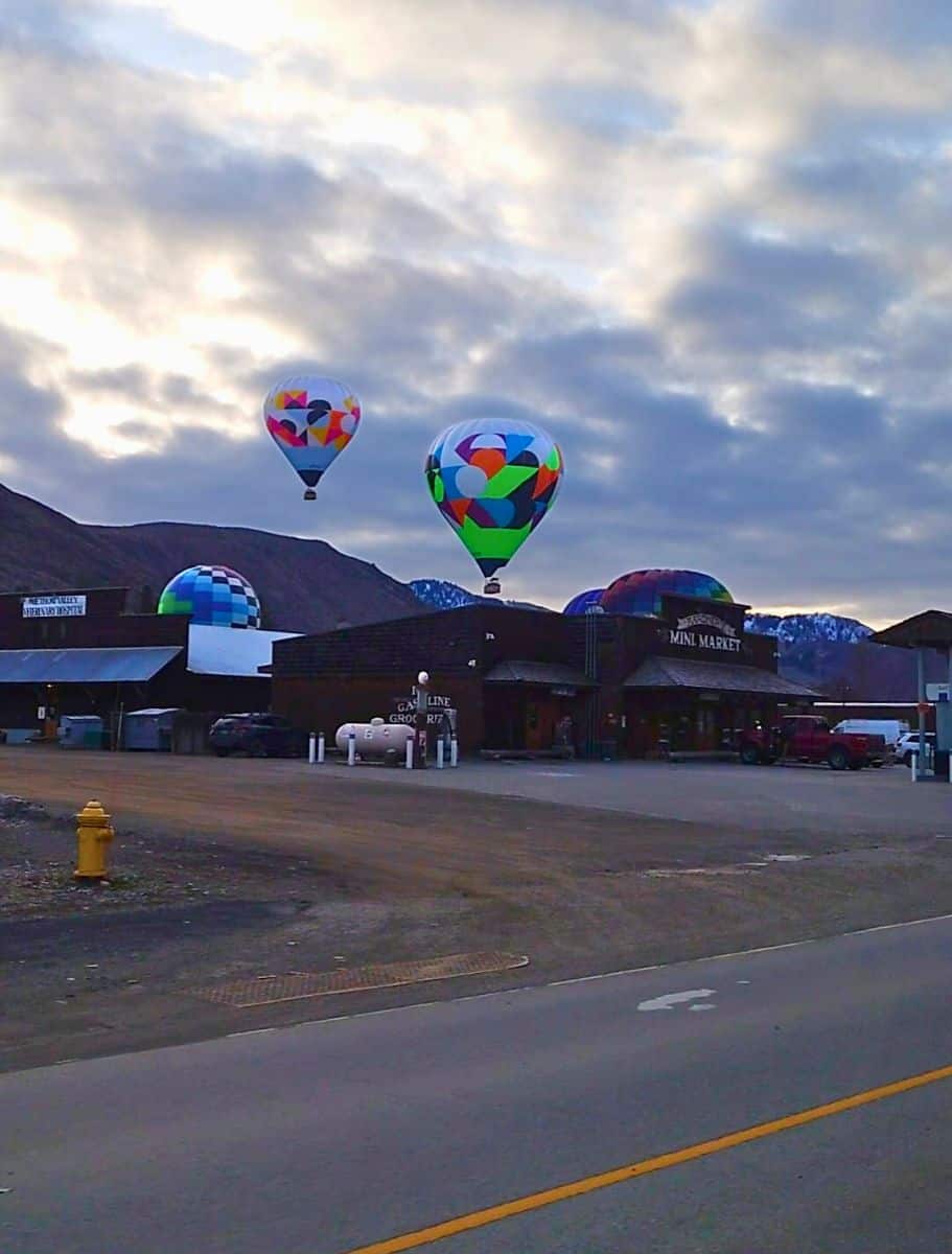 Hot air balloons rising in the distance behind quaint buildings in Winthrop, set against a mountainous backdrop and a cloudy sky.