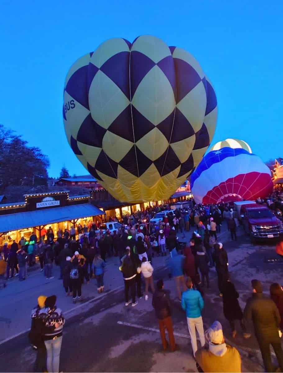 A majestic view of two giant hot air balloons lit up on a street in Winthrop as dusk falls, with a crowd of spectators enjoying the view