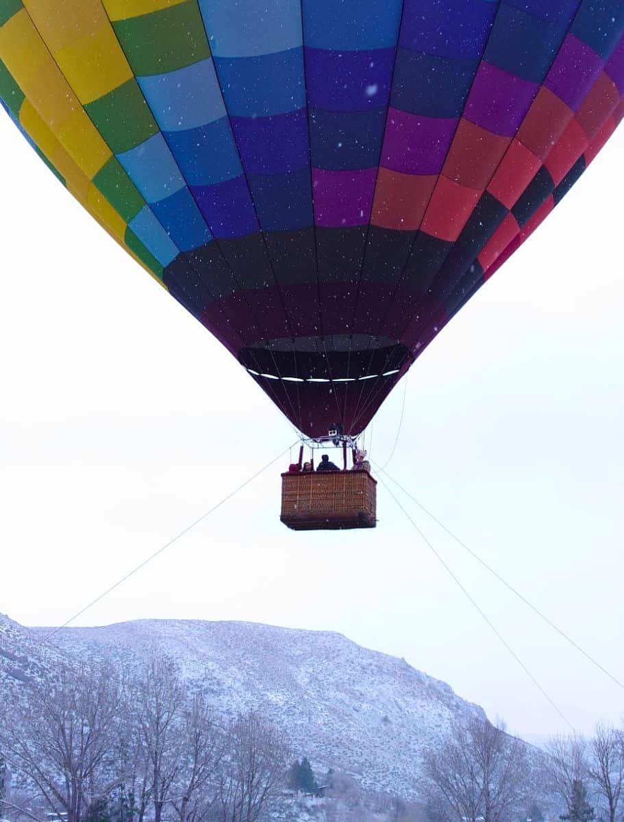 Close-up of a hot air balloon basket with passengers, set against the vibrant patchwork colors of the balloon and a snowy landscape below.