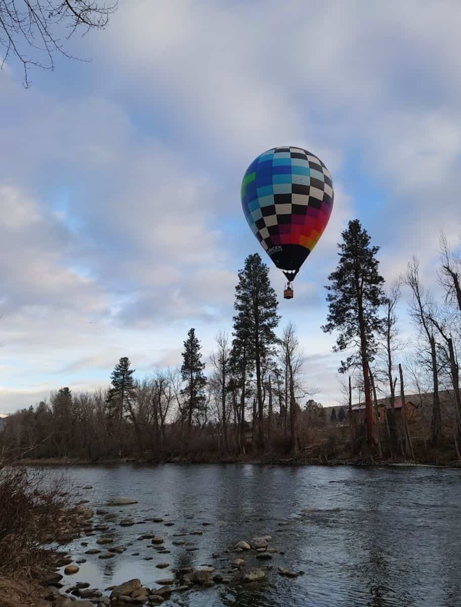 A checkered hot air balloon drifts above a serene river lined with trees, reflecting a peaceful setting
