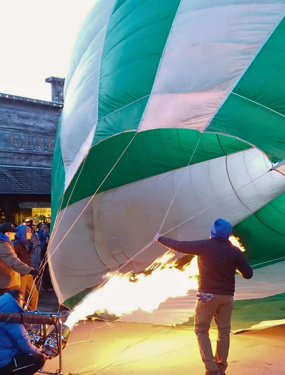 A hot air balloon in the process of inflating, with a person holding the envelope open as the burner flame fills it, set against an evening backdrop with onlookers.