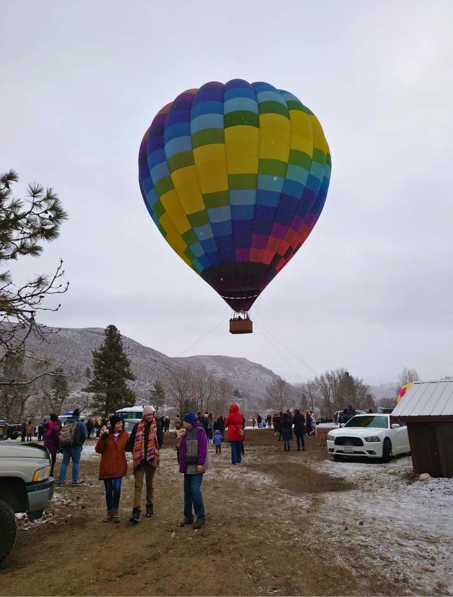 A fully inflated hot air balloon tethered to the ground, with a crowd of spectators around, under a grey, cloud-filled sky.