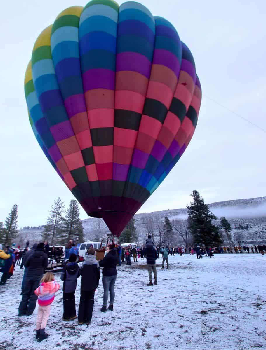A rainbow-colored hot air balloon stands tall in a field with a crowd of onlookers with snow on the ground