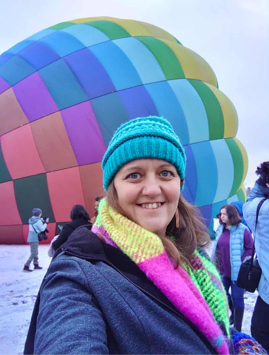 in a teal beanie and vibrant scarf poses in front of an inflating hot air balloon on its side on the ground