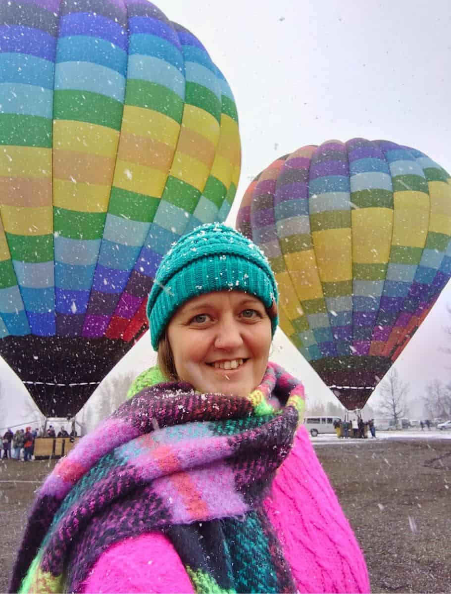 me in a teal beanie and colorful scarf smiles in the foreground, with two colorful hot air balloons in the snowy background