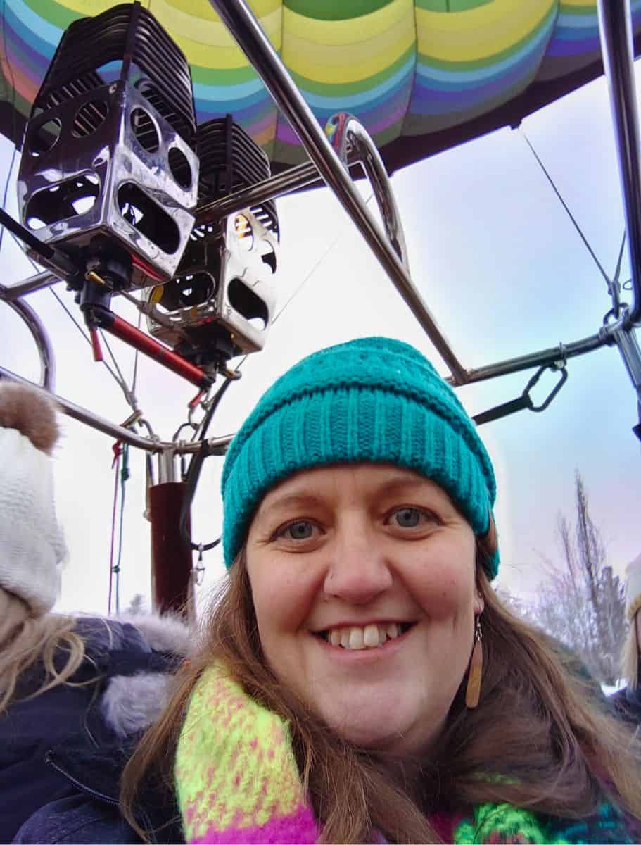 Me in a teal beanie and multicolored scarf inside a hot air balloon basket, with the balloon's burners visible overhead