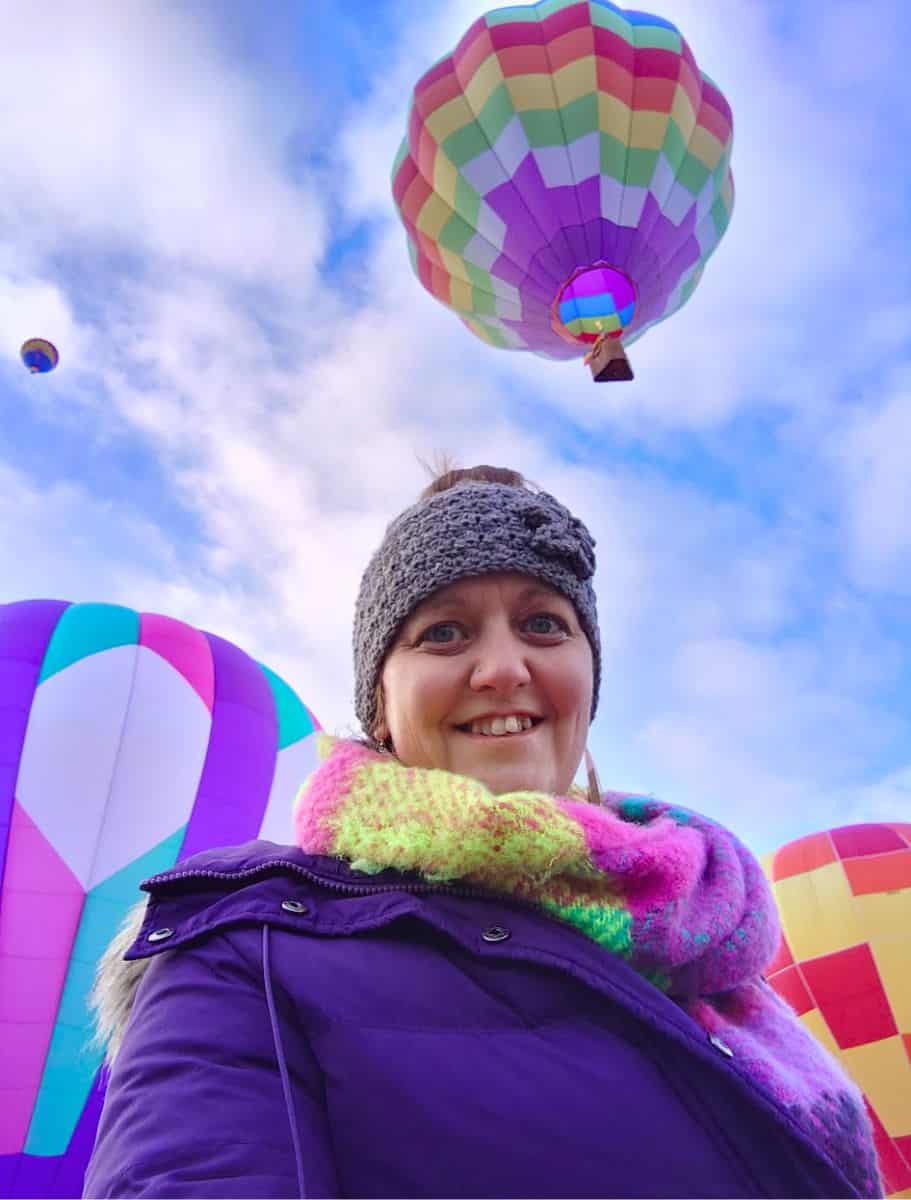 me in a purple jacket and colorful scarf smiles with large, inflated hot air balloons in the background, against a blue sky with soft clouds
