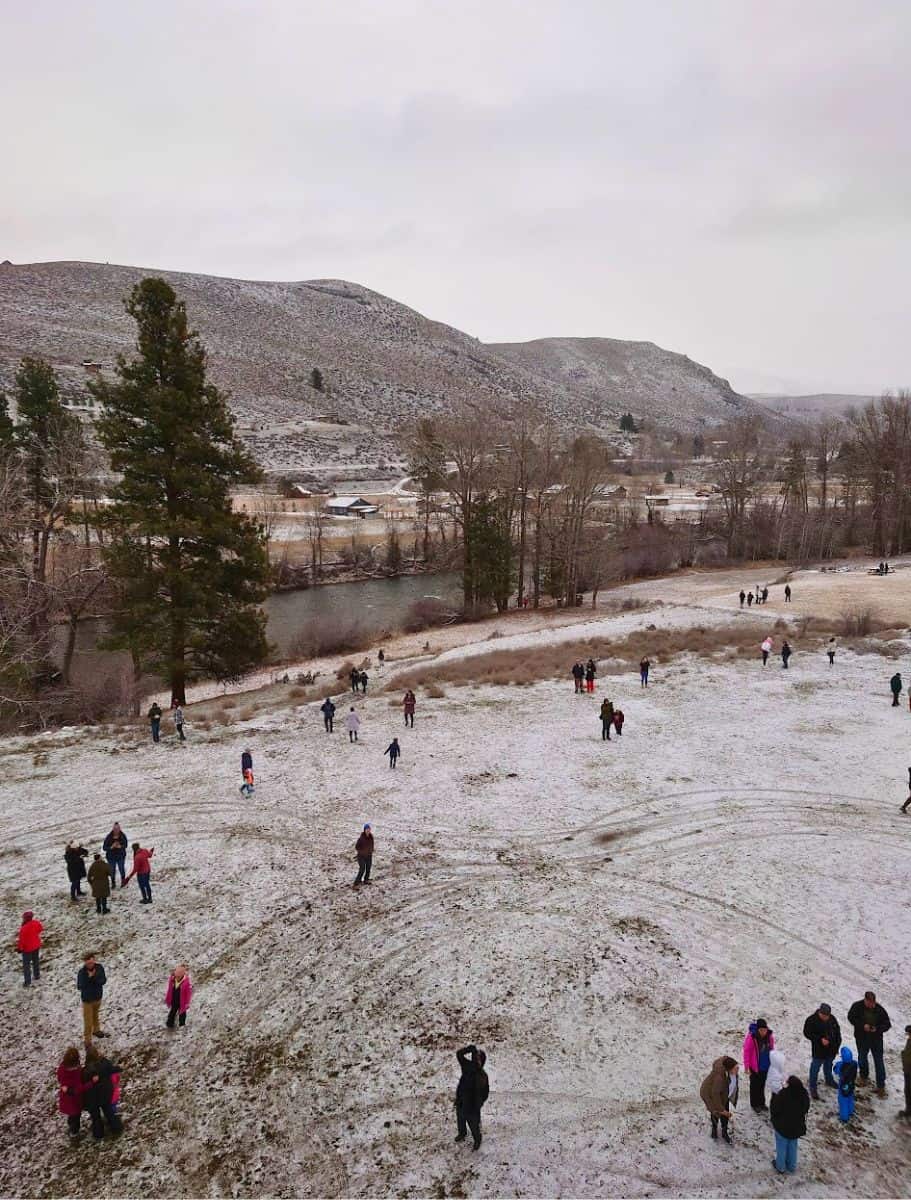 An aerial view from a hot air balloon shows people scattered across a snow-dusted field with a river and hills in the background
