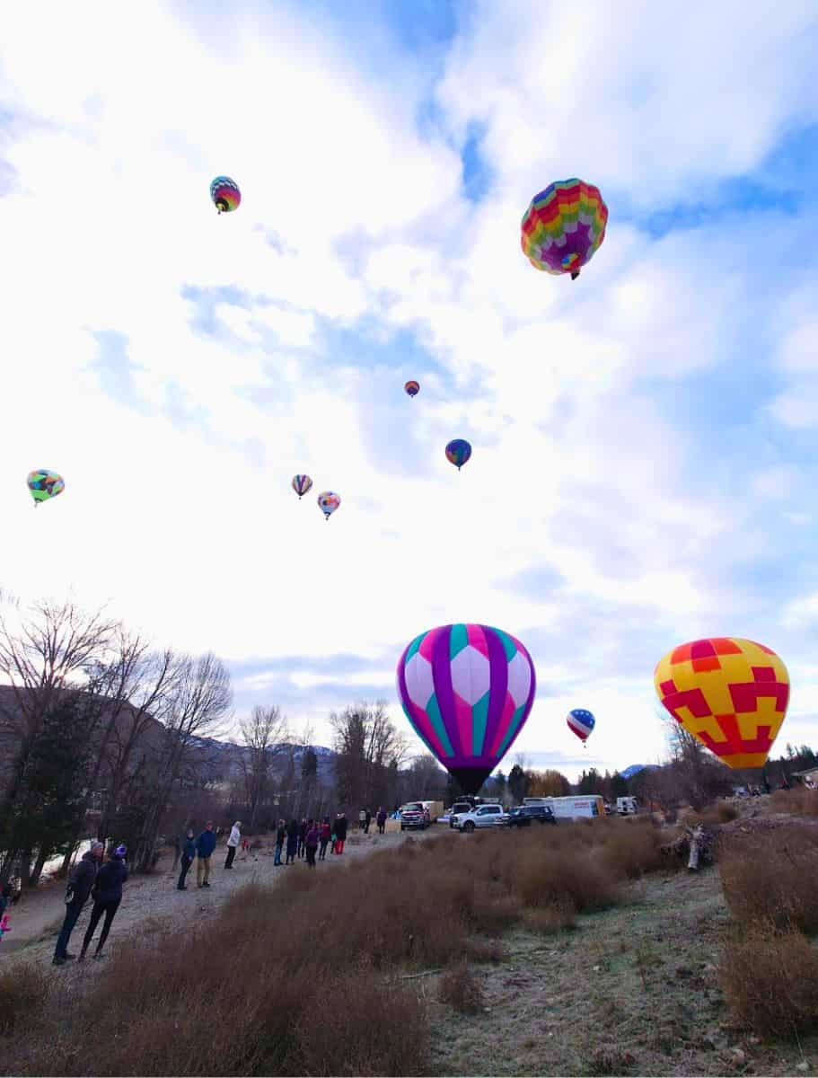A picturesque scene of multiple hot air balloons of various colors dotting the sky over a snow-speckled landscape with onlookers below