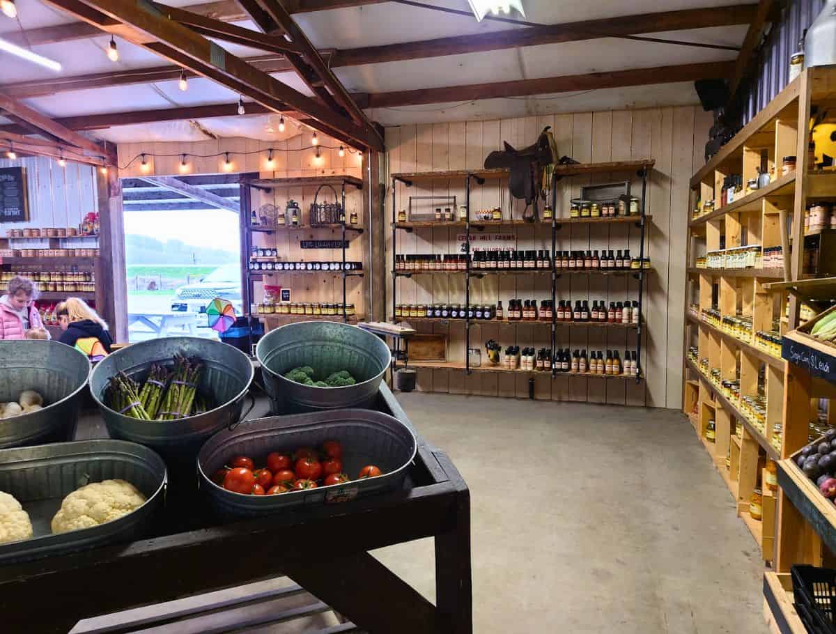 Rustic farm market with bins of fresh vegetables, wooden shelves stocked with local sauces and preserves, and string lights hanging from the ceiling. Visiting local farms and markets for fresh produce is one of the relaxing things to do in Oak Harbor for food lovers and families.