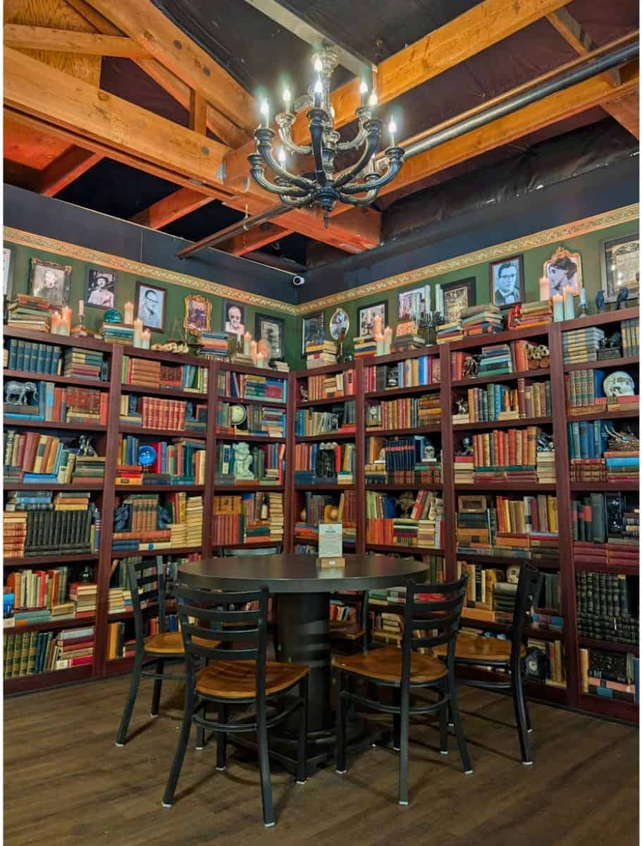 Cozy reading room with floor to ceiling bookshelves filled with vintage books, framed portraits, candles, and a chandelier hanging from exposed wooden beams. Relaxing in a unique bookstore or themed café is one of the cozy things to do in Oak Harbor for book lovers and rainy day visitors.