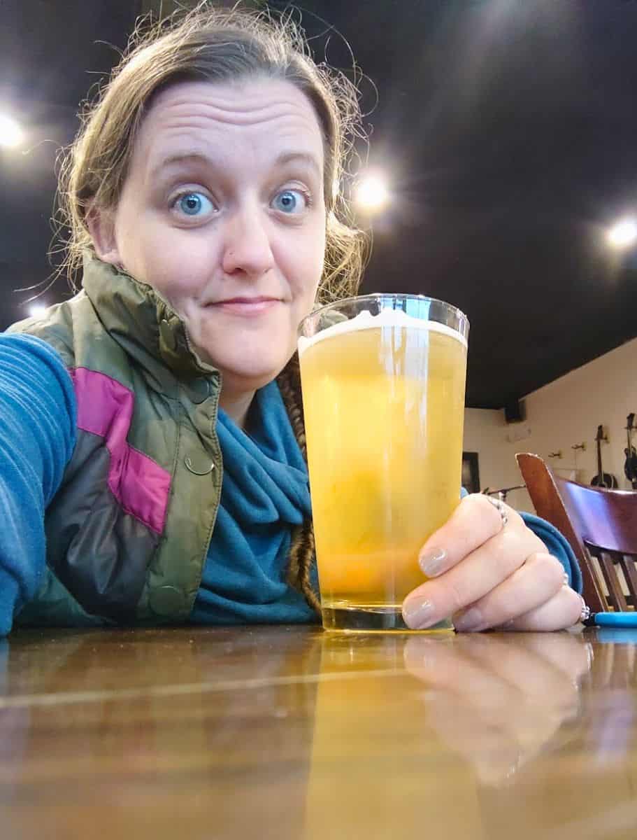 Woman smiling while holding a pint of light beer inside a casual brewery or taproom with warm lighting and wooden tables. Visiting local breweries and trying craft beer is one of the laid back things to do in Oak Harbor after a day of sightseeing.