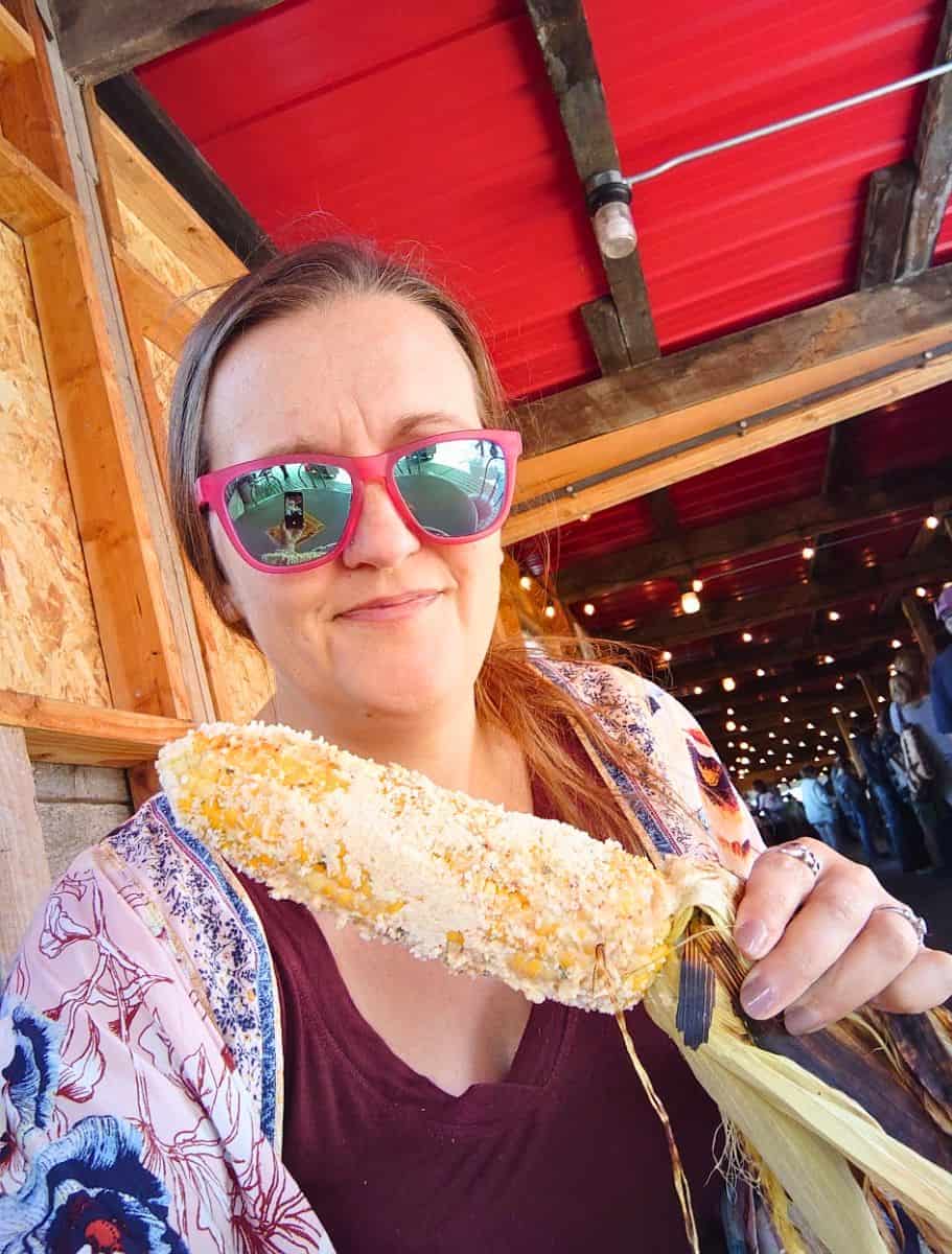 Woman wearing pink sunglasses holds a freshly roasted ear of corn covered in cheese while sitting inside a rustic farm stand with string lights overhead. Eating local farm food and fresh corn is one of the fun things to do in Oak Harbor during a summer visit.