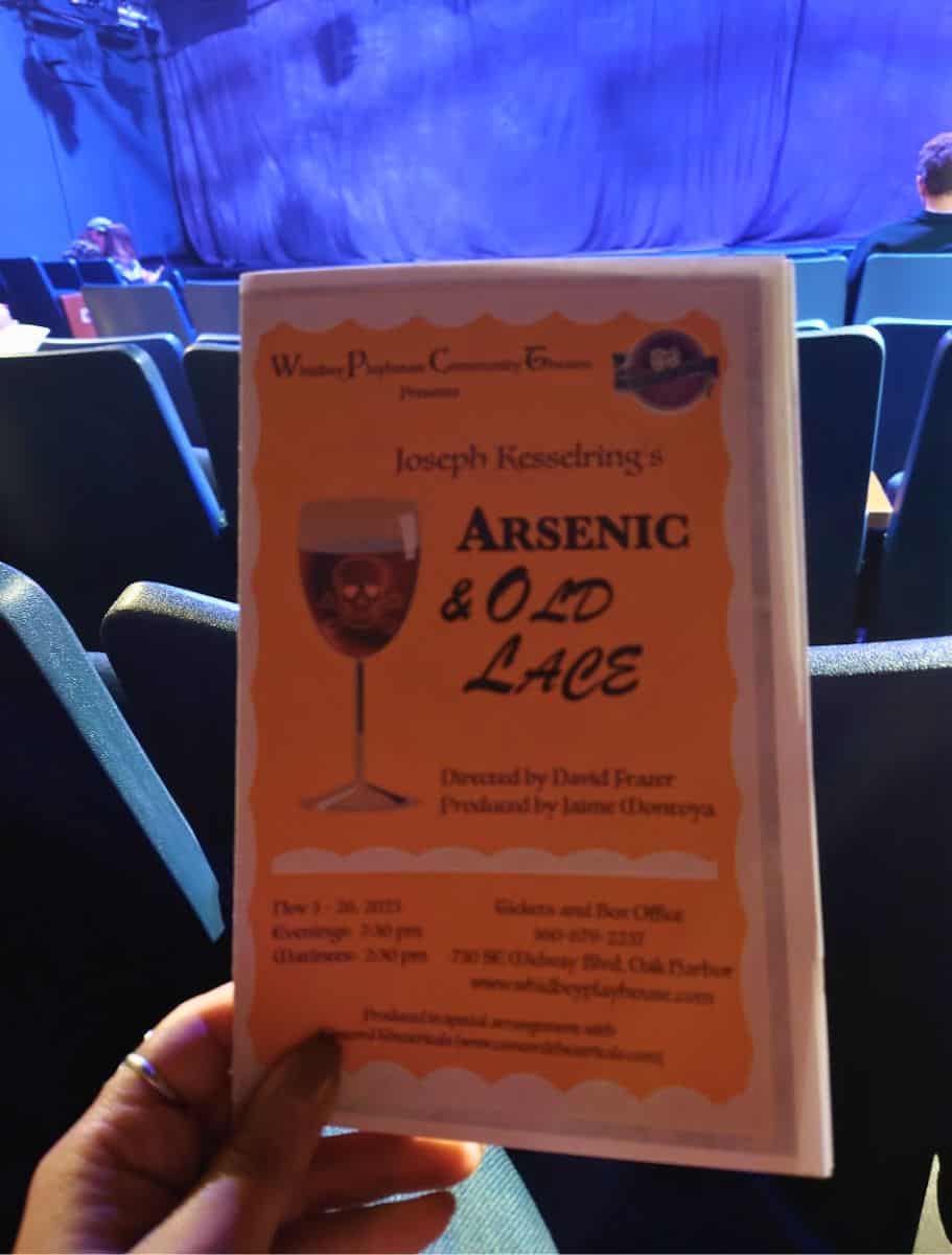 Person holding a playbill for "Arsenic & Old Lace" while seated inside a theater with a blue lit stage curtain in the background. Seeing a live performance at a local theater is one of the entertaining things to do in Oak Harbor during an evening out.